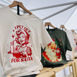 Beige t-shirt with red graphic and text 'I Put Out for Santa' on a wooden rack.